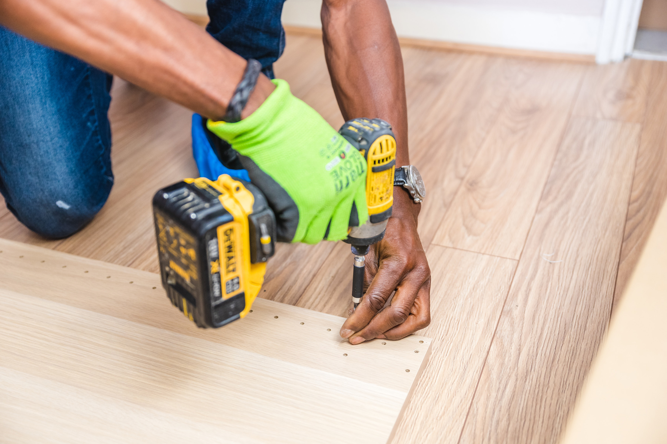 Close-up view of a BIPOC man's arms usign a Dewalt Impact Driver to assemble a cabinet.