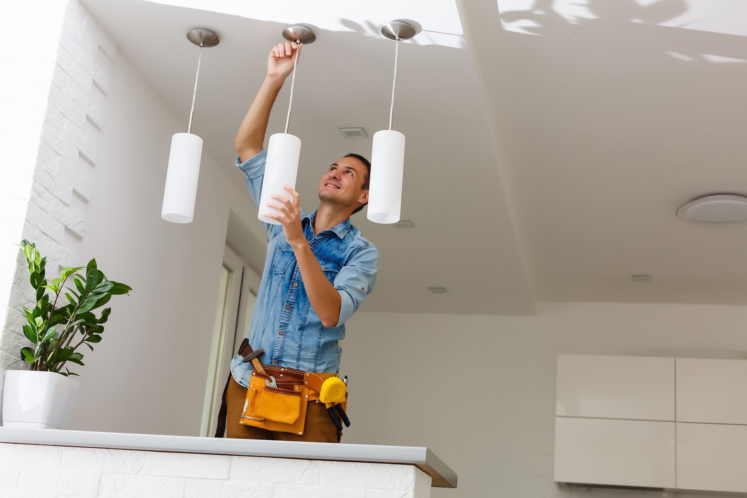 A man wearing a blue denim long sleeved shirt and leather work belt installing a white pendant light on the ceiling.