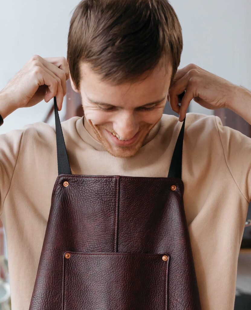 Smiling caucasian man with brown hair, wearing a tan t-shirt, putting on a dark brown leather apron.