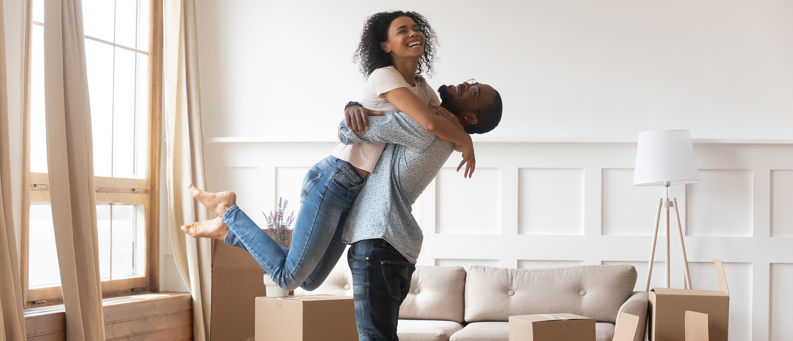 BIPOC couple in blue jeans hugging in a neutral coloured living room with moving boxes around them.