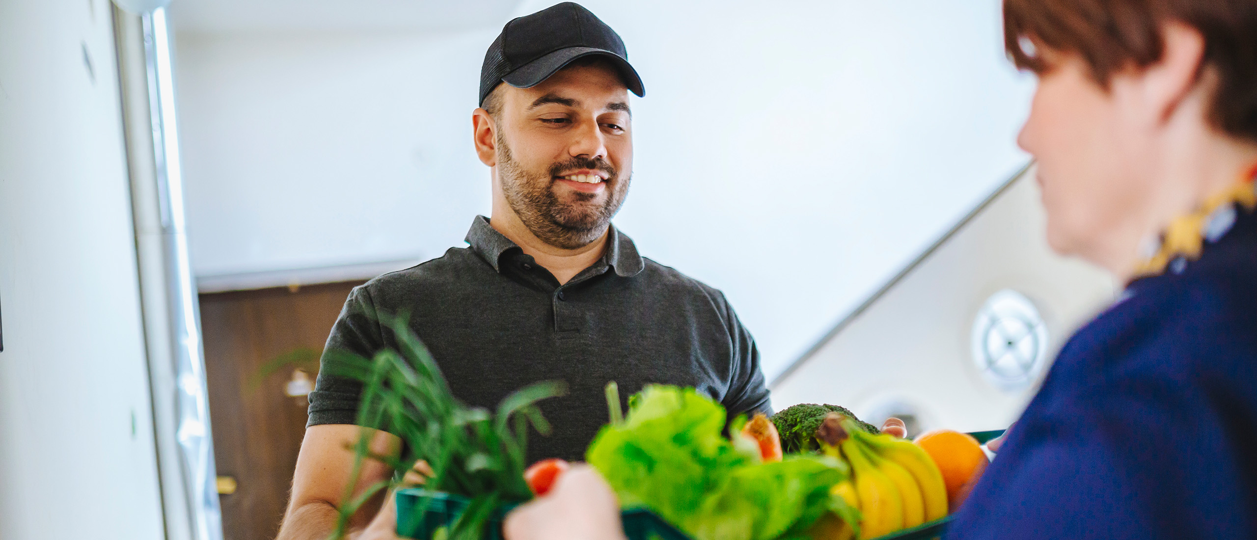 Middle aged man with facial hair wearing a black baseball hat and dark grey shirt delivering a large box of produce to a woman in a dark blue shirt.