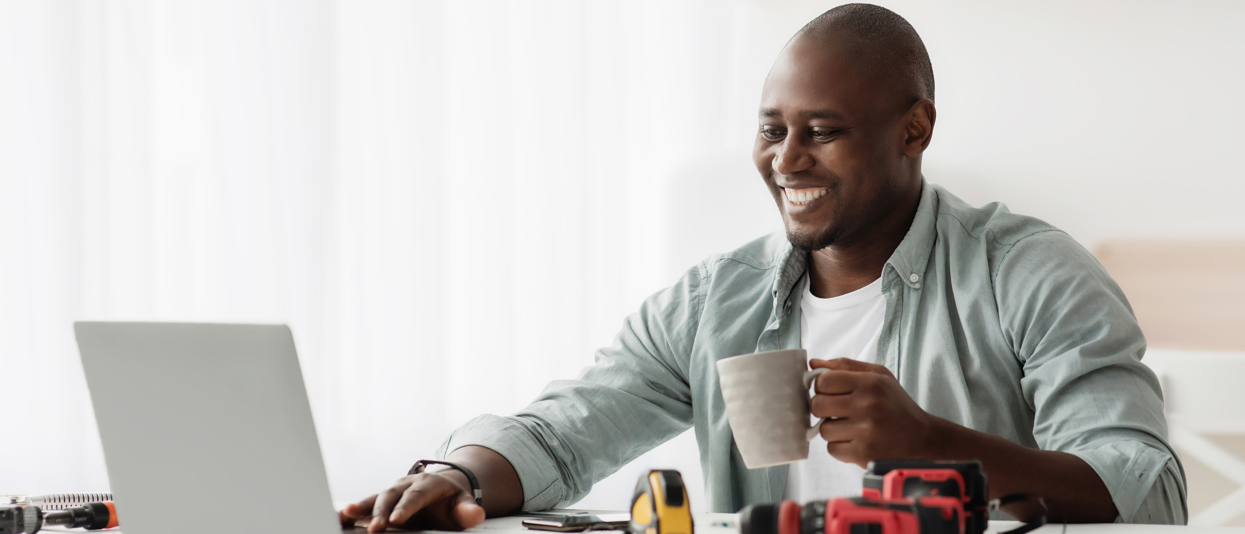 Smiling BIPOC man, wearing a white t-shirt under a light green long sleeve button-up, surrounded by tools, drinking a coffee while using his laptop computer.