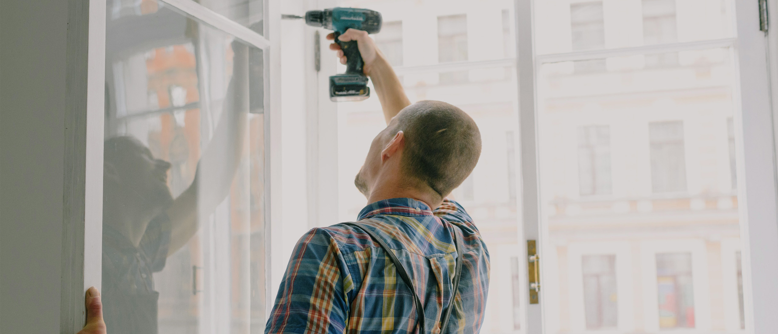 Caucasian man, in a colourful plaid short sleeve shirt, with a buzzed head using a Matika drill to secure a swining window's hinge.